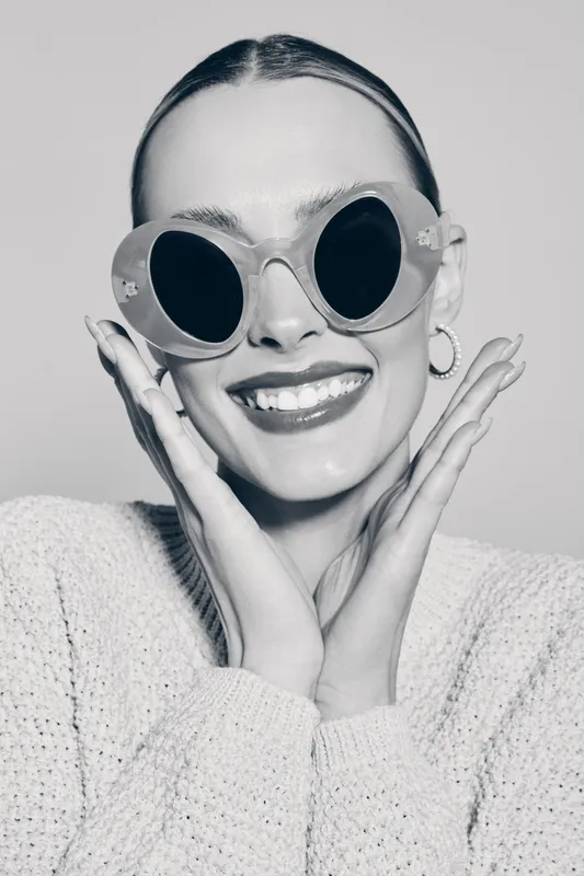 Happy Caucasian woman with light skin wearing large sunglasses and cozy sweater smiling with hands near face in studio. Concept of fashion, style, and joy.