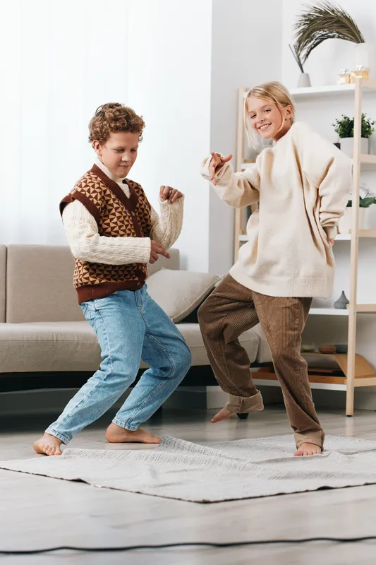 Two children dance joyfully in a bright living room, wearing knit sweaters and jeans, capturing a spontaneous moment of playful movement and carefree energy.
