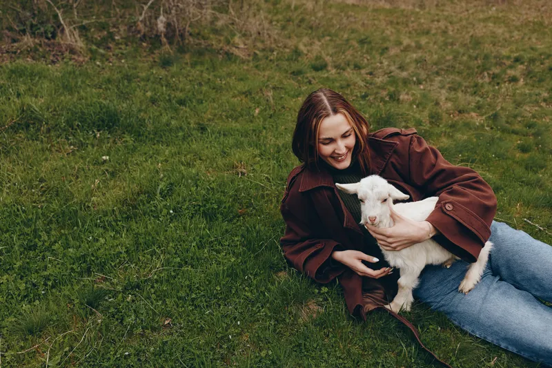 A woman lies on grass outdoors, holding a playful goat kid and sharing a gentle cuddle. Natural lighting enhances a warm, simple lifestyle moment with animals and nature.