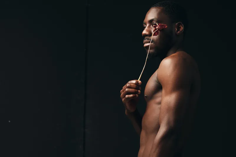 Black man with dark skin and muscular body holding dried flower near face in creative visual design style on dark background. Artistic portrait with minimalistic concept and studio lighting.