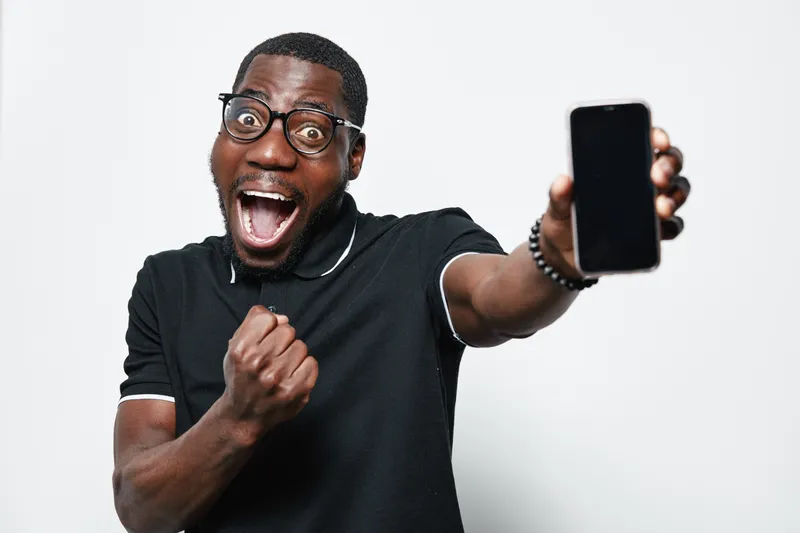 Excited African American man with dark skin and glasses holding smartphone showing blank screen, celebrating success with fist pump in casual black polo shirt on white background.