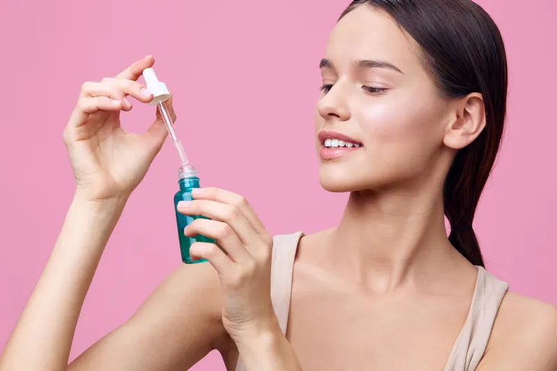 Young woman applying skincare serum, showcasing radiant skin with a pink background, highlighting beauty and self care in a fresh, modern style