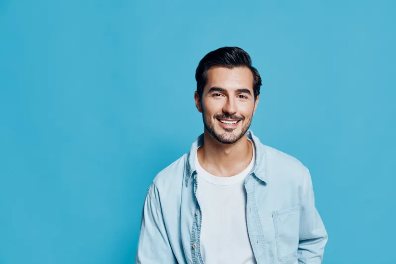 Smiling happy cheerful young man wear light blue shirt look at camera isolated on plain solid turquoise background studio portrait. People lifestyle concept