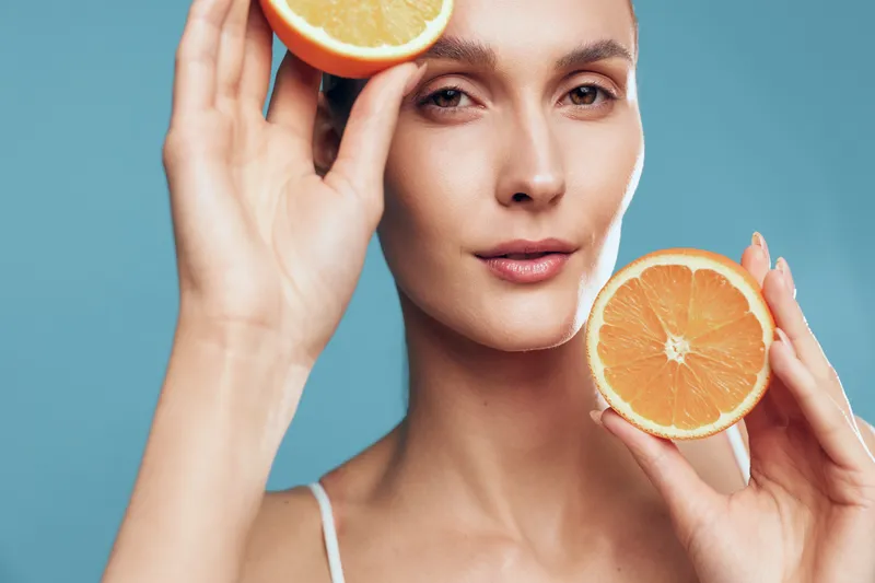Young woman with clear skin holds two halves of an orange near her face against a blue background, expressing freshness and natural beauty in a skincare concept.