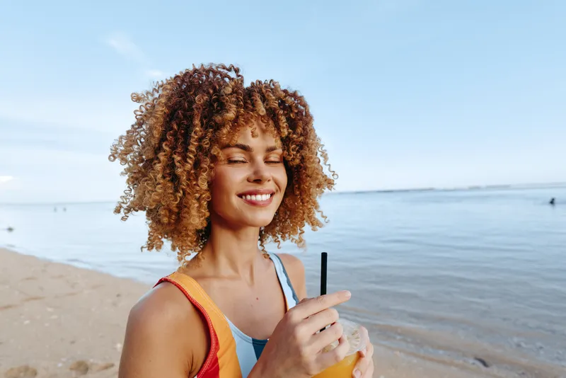 Young woman with curly hair smiles on a sunny beach while sipping a bright orange beverage, wearing a colorful tank top, enjoying a breezy seaside moment.