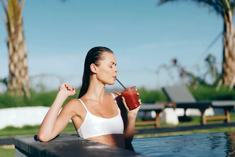 Young woman in white bikini drinking a red smoothie by the pool on a sunny day. Relaxed lifestyle concept with tropical background and clear blue sky for wellness and leisure.