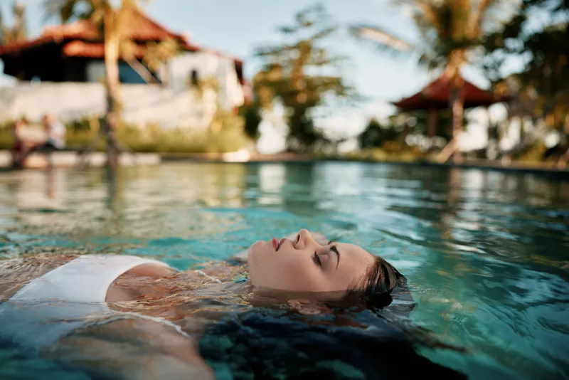 Young woman in white swimsuit relaxes floating on water surface of outdoor pool with tropical background and palm trees under sunlight during summer vacation.