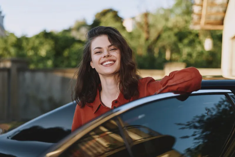 Young woman leans out of a sleek car window, smiling broadly as sunlight highlights her chestnut hair in a candid lifestyle moment during golden hour as a breeze lifts her jacket and the urban