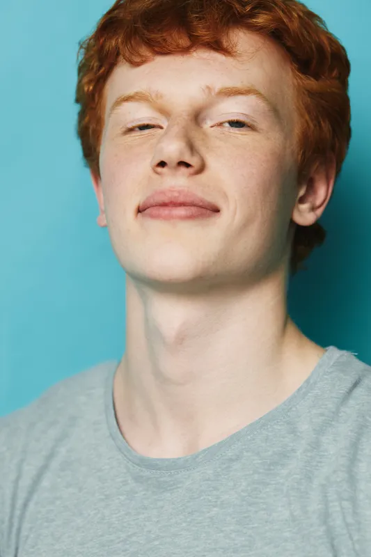Portrait of young man with red hair and freckles wearing grey t shirt against blue background. Confident expression and casual style with natural skin and youthful look.