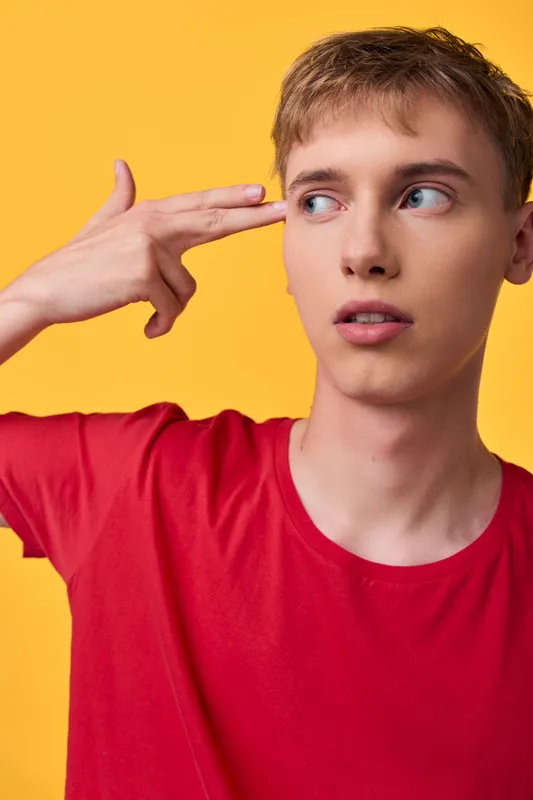 Gesture of a thoughtful youth with finger gun toward temple, red shirt against a bright yellow backdrop, conveying curiosity, hesitation, or playful thinking in a modern style