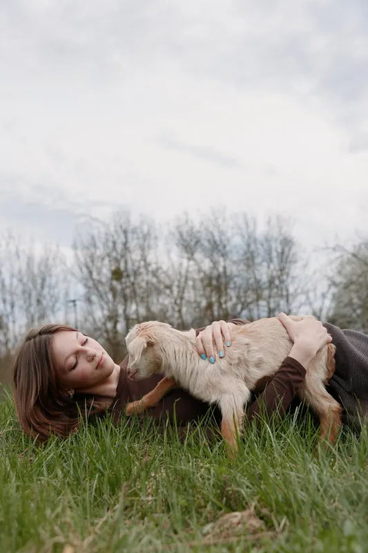 Young woman with brunette hair lies on the grass gently holding a small goat in a natural outdoor setting during a calm spring day.