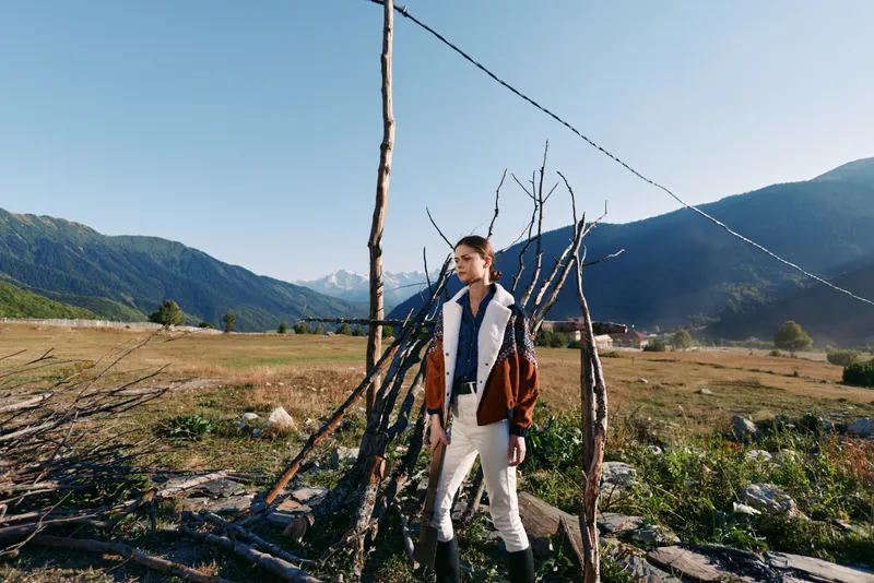 Woman in jacket and boots stands in a field with mountains behind, portrait style shot in nature and countryside. Rustic wooden structure and clear sky create serene travel mood.
