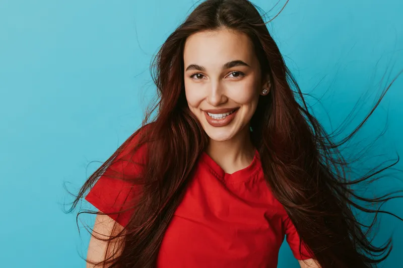 Young woman with long hair wearing red shirt smiling and looking at camera on blue background. Portrait of happy female with natural makeup and joyful expression in studio lighting.