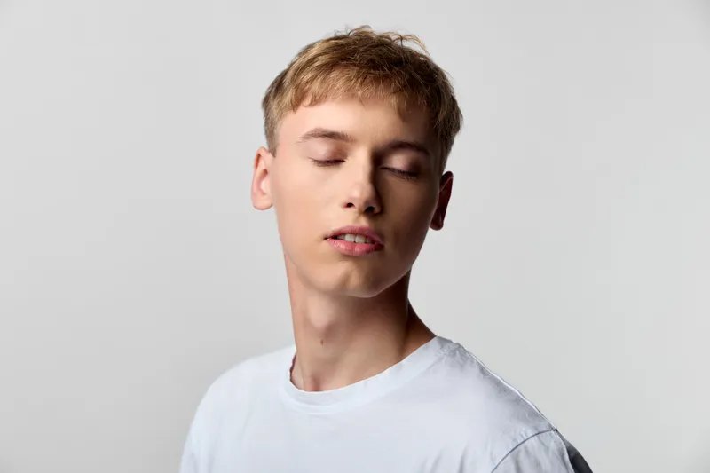 Young man with closed eyes wearing a white tshirt in a calm studio setting, a serene fashion pose that conveys relaxed mood, minimal light and quiet confidence in a simple candid style