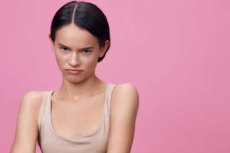 Young woman with a neutral expression in a beige top against a pink background, showcasing modern fashion and emotional depth for lifestyle imagery