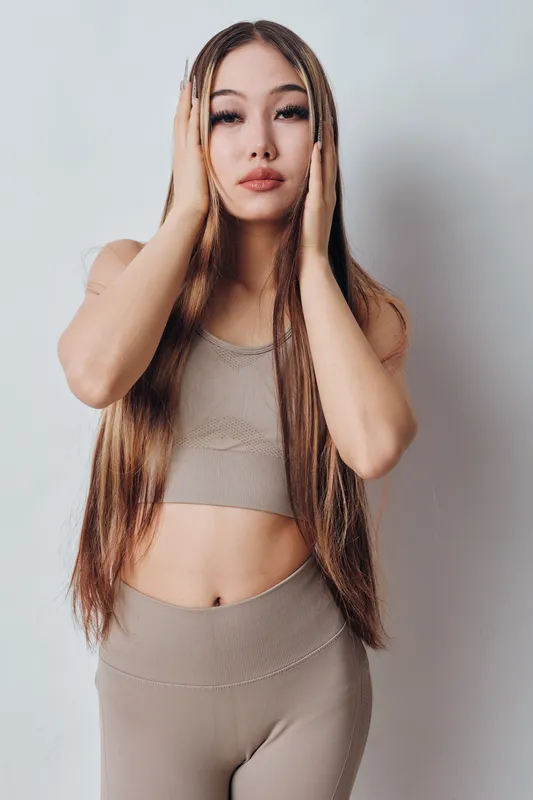 Young woman with long straight hair wearing neutral workout clothes standing and touching face against white studio background for beauty and fitness concept.