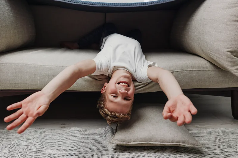 Playful boy with curly hair lies upside down between sofa cushions, smiling with arms stretched out in cozy living room. Concept of childhood joy, relaxation, and home comfort.