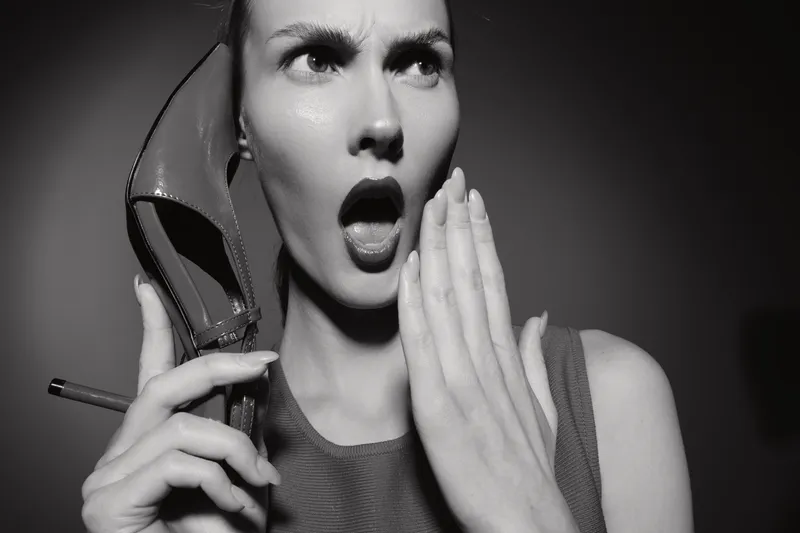Surprised white woman with light skin holds high heel shoe and cigarette near face with open mouth in black and white photo.
