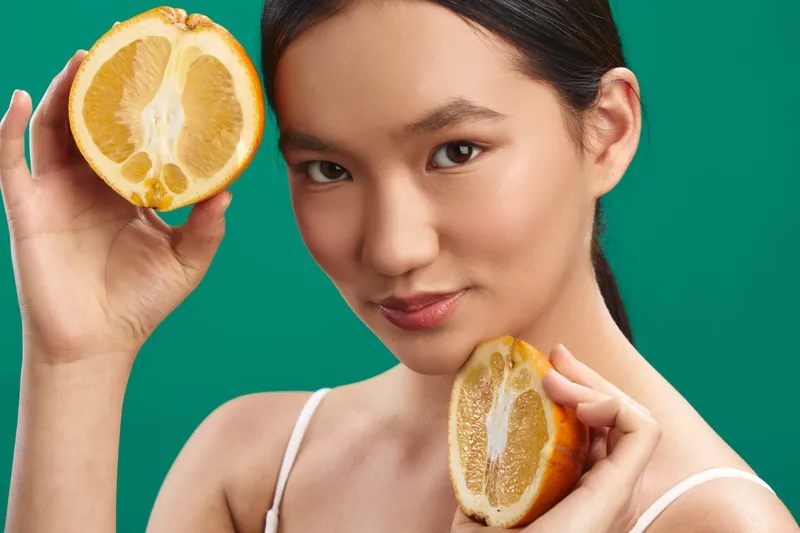 asian woman holds lemon halves near her face in a bright studio, creating a fresh, vibrant portrait with citrus accents and a playful, stylish mood against a green backdrop.