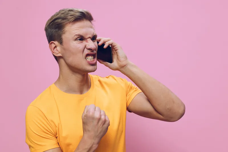Angry young man with short hair wearing yellow t-shirt holding mobile phone angry yelling or shouting isolated on pink background studio portrait. People emotion concept