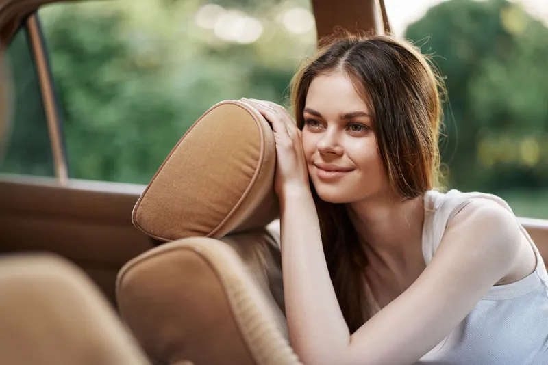 Young woman smiling softly in cozy car interior, showcasing relaxed emotions and natural beauty in a lush green background, ideal for lifestyle and travel themes.