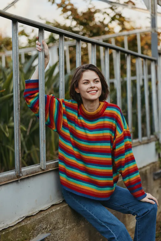 Young woman sweater striped colorful smile casual outdoors leaning fence portrait, cozy knit and relaxed pose with natural light in urban garden setting for lifestyle photography