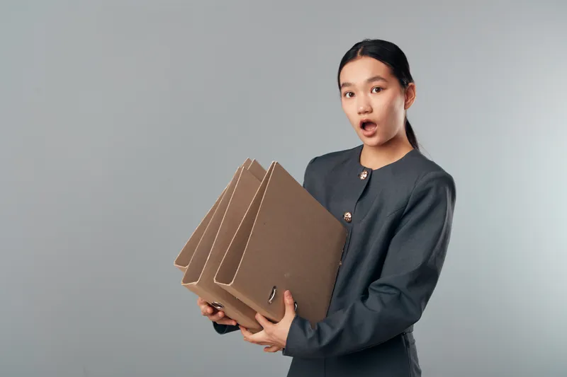Young woman in dark clothing holding several large cardboard boxes with surprised expression on face against gray background, isolated studio portrait of delivery or moving concept.