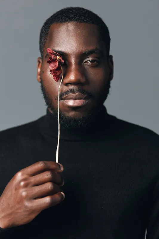 Creative conceptual portrait of African man holding dried flower in front of eye with serious expression in studio. Visual style with dark skin and artistic mood on gray background.