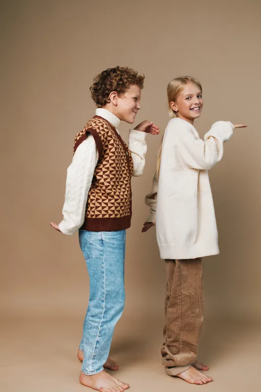 Two playful children stand back to back, smiling and ready for adventure in cozy retro outfits, capturing carefree friendship, imagination, and soft, timeless charm in a warm studio.