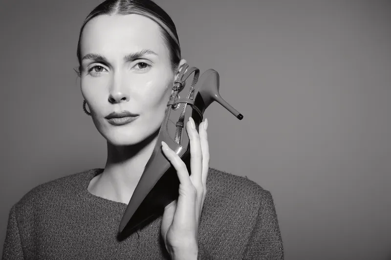 Elegant white woman with light skin holds black high heel shoe near face, wearing knit sweater, monochrome studio portrait, fashion concept.