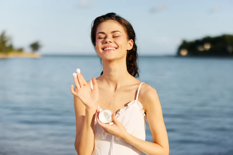 Young woman at the seaside gently applying sunscreen, smiling with closed eyes, wearing a light dress, standing near calm blue water with distant shoreline in the background.