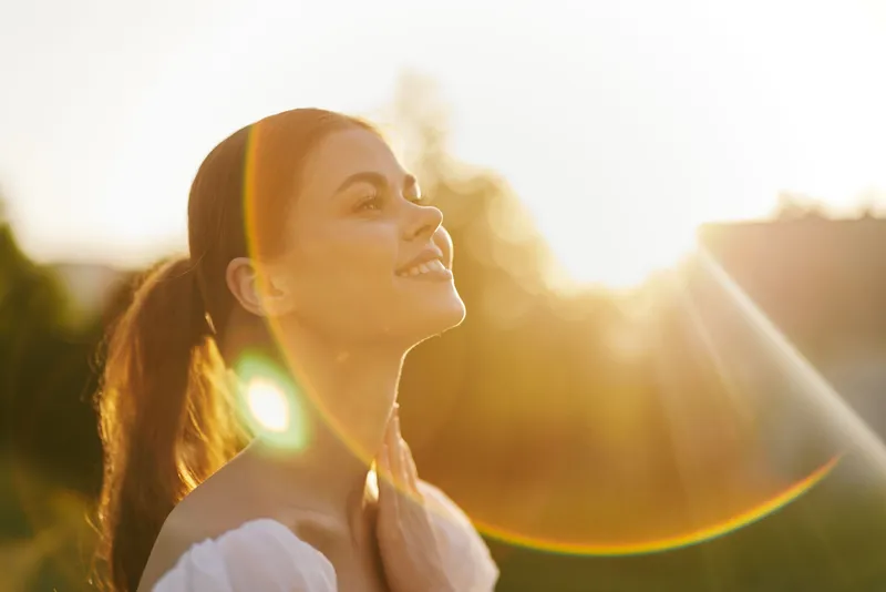 Young woman enjoying sunlight outdoors with a joyful expression, wearing a white off shoulder dress surrounded by nature, showcasing a sense of serenity in summer
