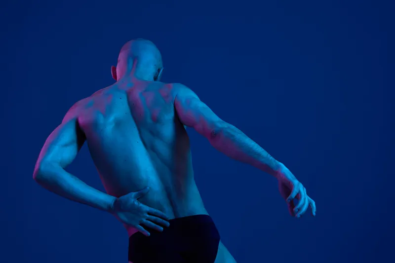 Athletic man posing against a vibrant blue background, highlighting his muscular back and strong physique in a dramatic light setup. This image captures strength and artistry.
