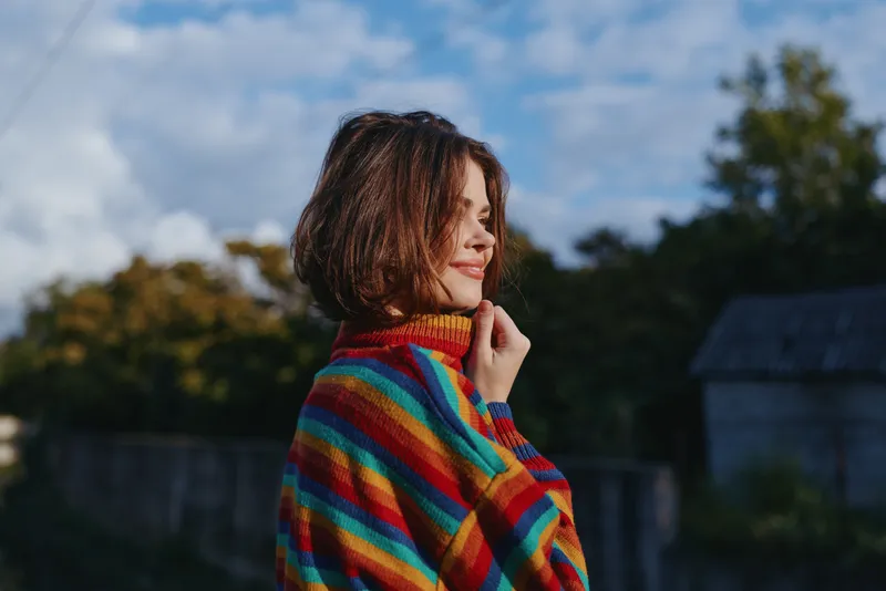 Woman, striped sweater, smiling, outdoor, young adult, short hair, portrait, countryside — 22-year-old casual model leaning slightly with hand to chin in colorful knit, soft sunlight, lifestyle