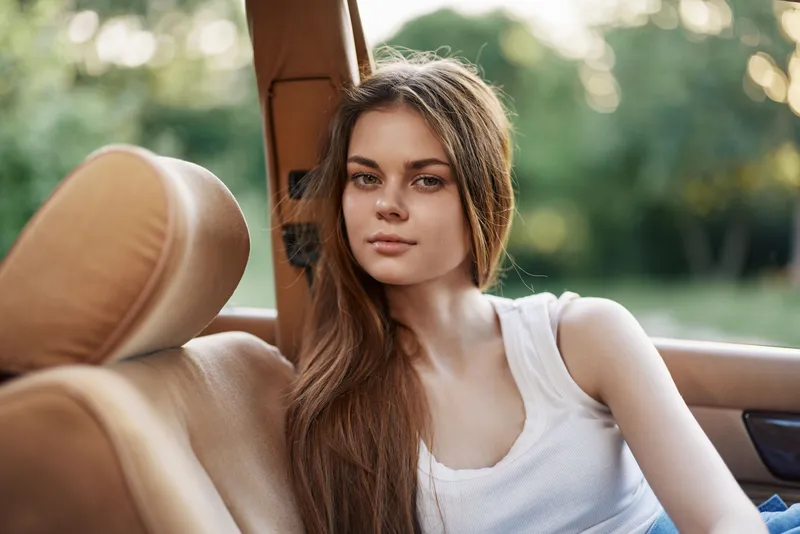 Young woman with long hair in a white tank top sitting in a convertible car, showcasing a serene expression against a blurred green background.