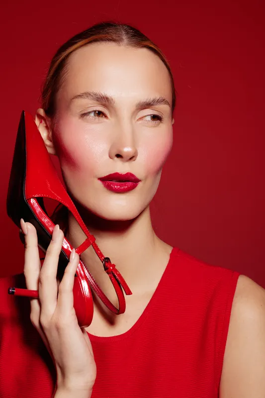 Woman in red dress holds red high heel shoe near face against red background. Fashion concept with bold makeup and stylish accessories in studio lighting.