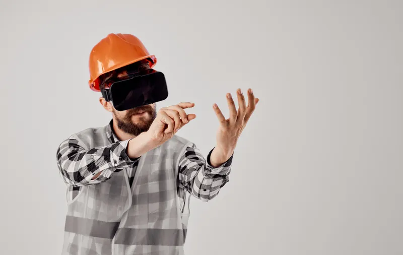 Инженер-строитель в оранжевой каске, клетчатой рубашке, 3D-очках, жестикулирует руками