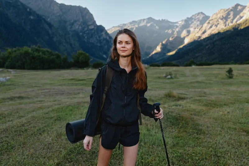 A cheerful hiker stands in a wide meadow with towering mountains in the background. She wears a black jacket and shorts, carrying a mat and trekking pole, ready for adventure.