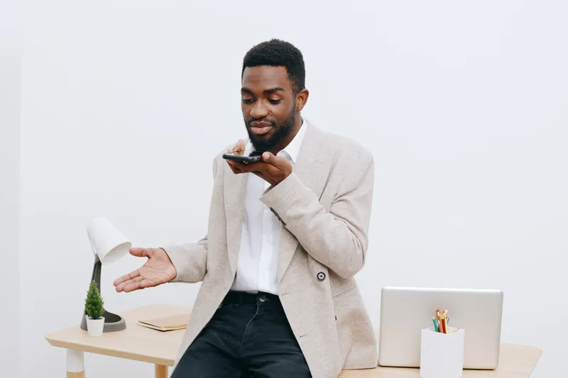 Professional young man with dark skin and short hair using smartphone while standing near a desk in modern office interior. Casual beige blazer over white shirt, confident expression, minimalistic wor