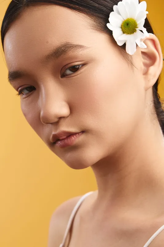 Asian woman gazes softly toward the camera, with a delicate flower tucked behind her ear. She wears light makeup and a gentle smile, creating a serene portrait that exudes quiet confidence and