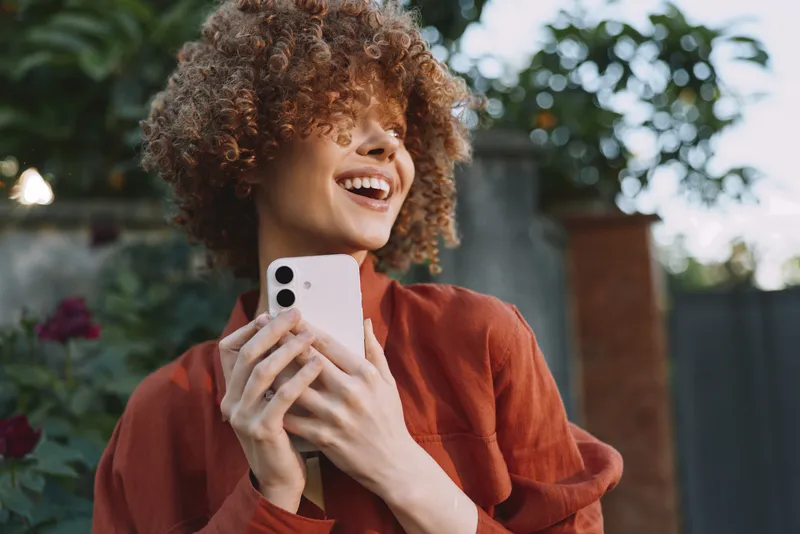 Smiling woman with curly hair holding a phone outdoors, demonstrating lifestyle and connection with nature through warm, natural light