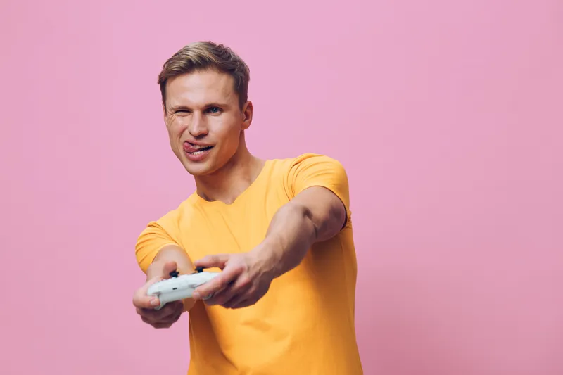 Smiling young man with short brown hair wearing yellow t-shirt holding game controller, looking at screen, isolated on pink background, indoor studio portrait, people lifestyle concept, joy, entertain