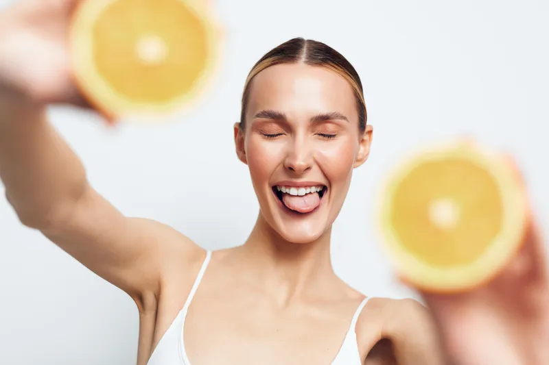 Woman laughing citrus orange fresh studio skincare cheerful white top, eyes closed, raised arm, blurred orange slices background, joyful beauty portrait