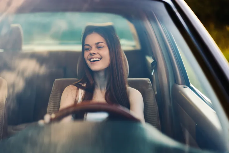 Young woman smiling while driving a car, enjoying freedom, in a sunny environment, representing joy and adventure in a casual setting.