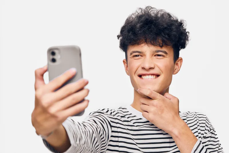 smiling man taking selfie with smartphone, wearing striped shirt, curly hair, casual look, bright background, youthful expression, indoor portrait.