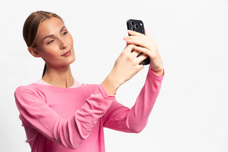 Young woman in pink sweater taking a selfie with black smartphone on white background. Concept of social media, technology, and self-portrait photography in studio lighting.