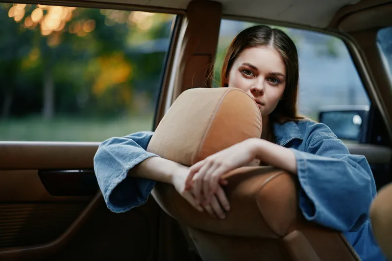 Young woman in denim jacket resting on car seat, with calm expression, capturing a nostalgic moment in a natural outdoor background during sunset.