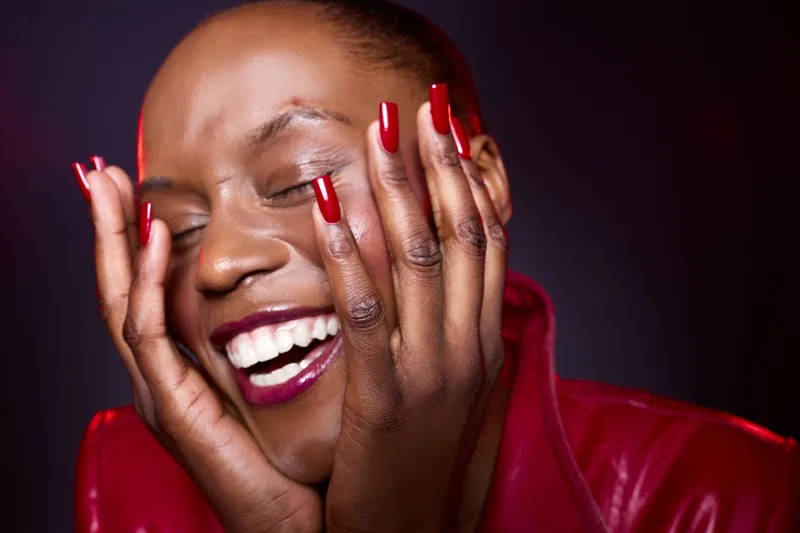 Close-up of happy dark skinned African woman with red nails and lipstick smiling with closed eyes and hands on face. Concept of joy, beauty, fashion, and self-expression in studio lighting.