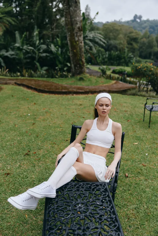 Young woman in stylish white activewear relaxing outdoors, showcasing a healthy lifestyle and wellness concept in a lush green garden