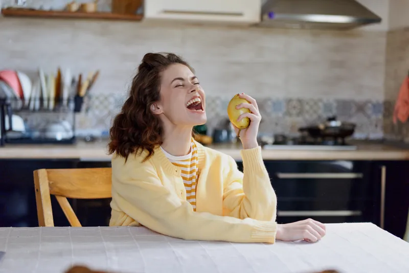 Young woman in a yellow sweater sits at a bright kitchen table, laughing with an apple in hand, capturing a warm, relaxed home moment full of casual joy.